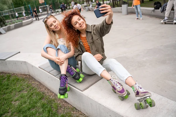 Cheerful friends in rollers skates taking selfie on border bench in skate park — Stock Photo