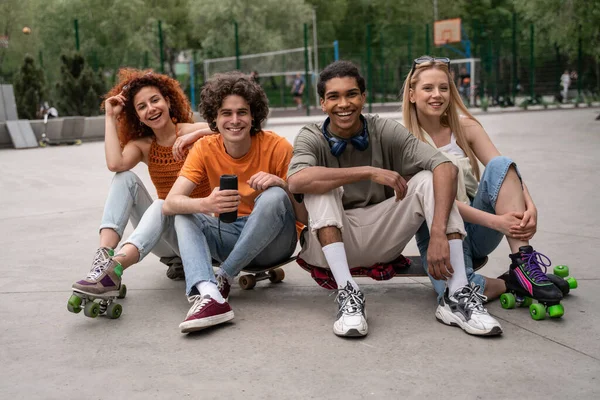 Patinadores multiétnicos felices sentados sobre asfalto en skate park con altavoz de música portátil - foto de stock