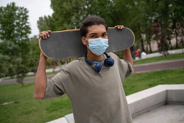 Молодой африканский американец в медицинской маске держит скейтборд за шеей и смотрит в камеру — стоковое фото