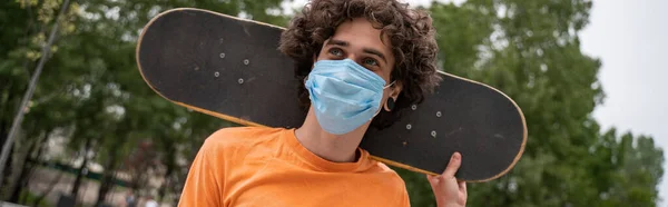 Кучерявий чоловік в медичній масці тримає скейтборд за шиєю, банер — стокове фото