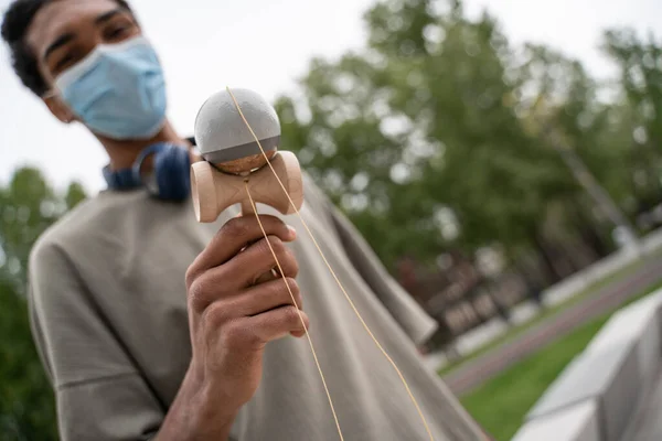 Borrosa africano americano hombre en médico máscara jugando kendama juego al aire libre - foto de stock