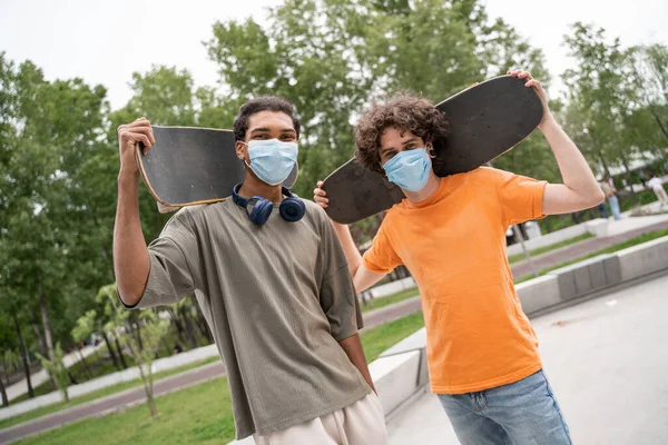 Amigos multiétnicos en máscaras de seguridad mirando a la cámara mientras sostiene patines - foto de stock