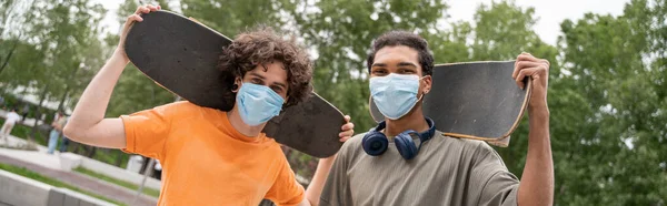 Inerracial skaters in medical masks looking at camera outdoors, banner — Stock Photo