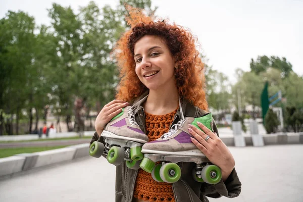 Cheerful redhead woman with roller skates on neck smiling at camera outdoors — Stock Photo