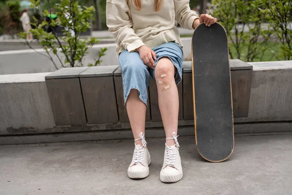 Cropped view of skater with adhesive plasters on injured knee sitting on border bench — Stock Photo