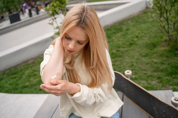 Mulher loira aplicando gesso adesivo no cotovelo ferido perto de skate no banco — Fotografia de Stock