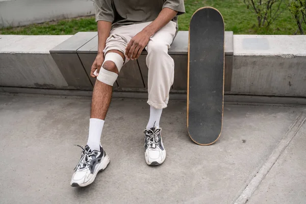 Cropped view of african american man bandaging injured knee near skateboard — Stock Photo