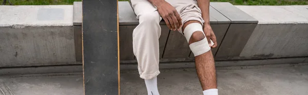 Cropped view of african american skater bandaging injured knee while sitting on border bench, banner — Stock Photo