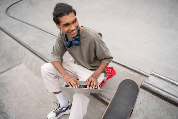 Vue grand angle de joyeux étudiant afro-américain utilisant un ordinateur portable à la frontière près de skateboard — Photo de stock