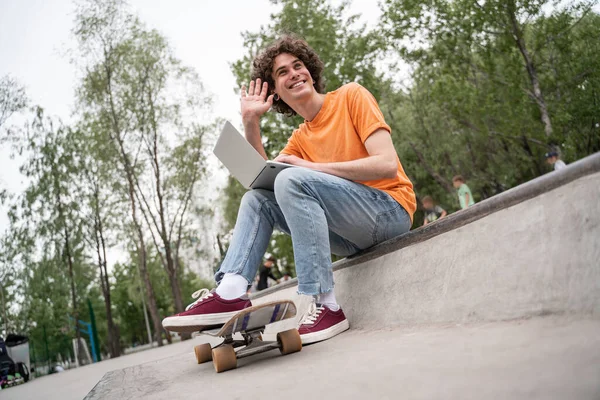 Cheerful skateboarder with laptop waving hand while sitting outdoors — Stock Photo