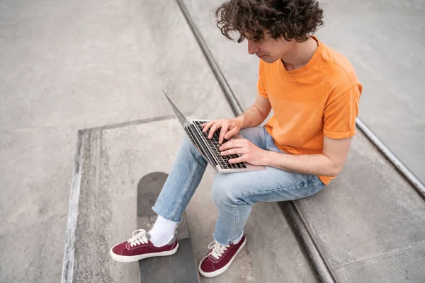 Young curly skateboarder typing on laptop in skate park — Stock Photo