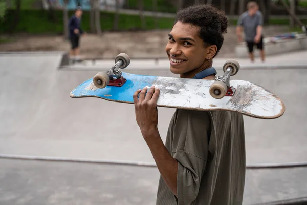 Jovem afro-americano homem sorrindo para a câmera enquanto segurando skate no ombro — Fotografia de Stock