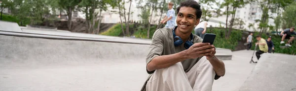 Hombre afroamericano feliz con auriculares y teléfono móvil sentado en el parque de skate, pancarta - foto de stock