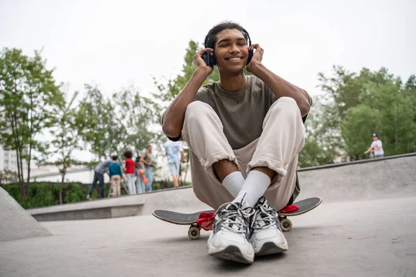 Homme afro-américain joyeux écouter de la musique dans les écouteurs tout en étant assis sur le skateboard — Photo de stock