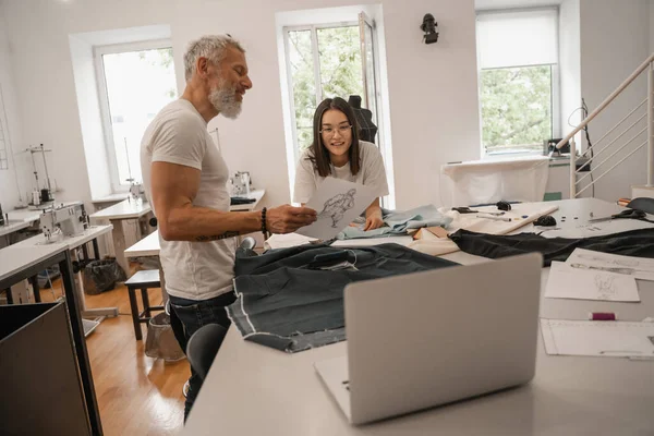 Lächelnde multiethnische Designer betrachten Skizze in der Nähe von Stoff und Laptop — Stockfoto