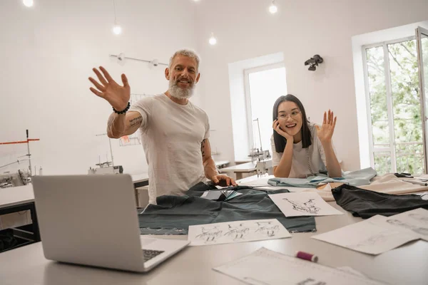 Sonrientes diseñadores interracial agitando las manos en el portátil cerca de bocetos y tela - foto de stock