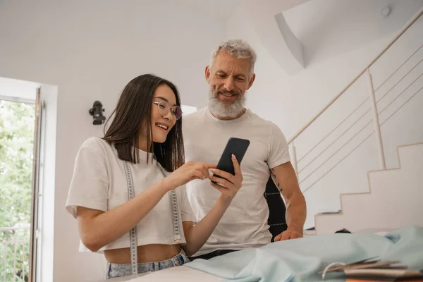 Sonriente asiático diseñador holding smartphone cerca de colega en taller - foto de stock