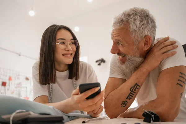 Sonriente asiático diseñador holding smartphone cerca de colega en taller - foto de stock