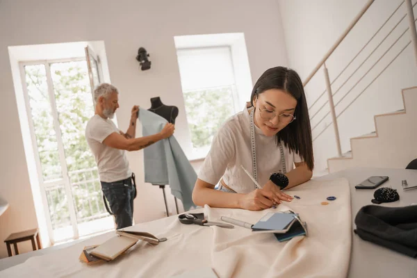 Asian designer drawing on fabric near blurred colleague in studio — Stock Photo