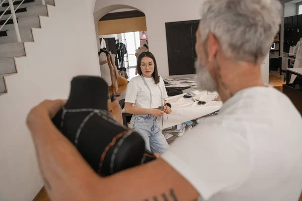 Asian designer looking at blurred colleague with mannequin — Stock Photo