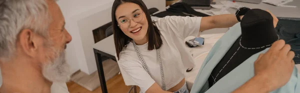 Lächelnder asiatischer Designer schaut Kollegin in der Nähe von Schaufensterpuppe an, Banner — Stockfoto
