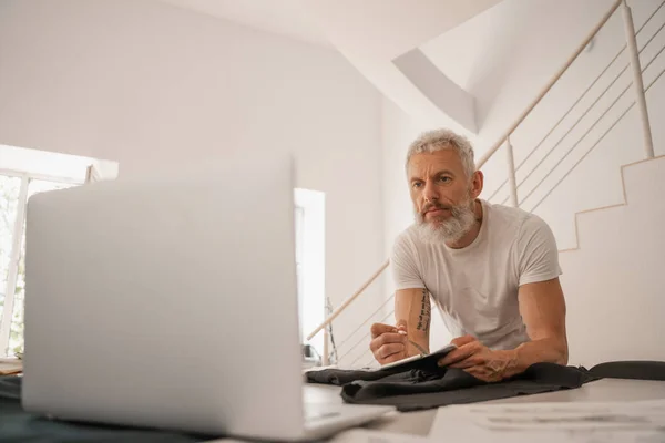 Mature designer looking at laptop near blurred cloth — Stock Photo