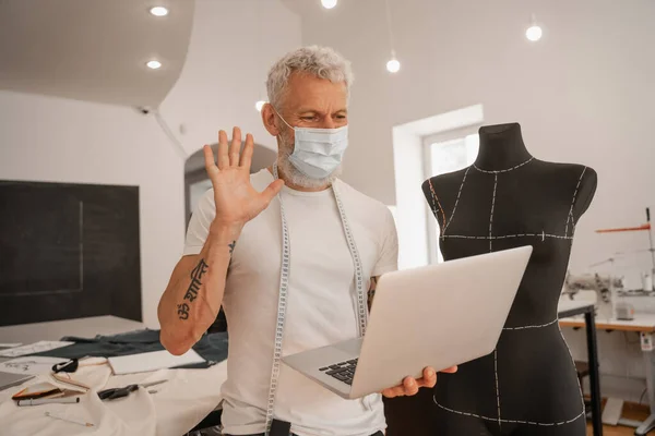 Designer in medical mask waving hand at laptop in workshop — Stock Photo