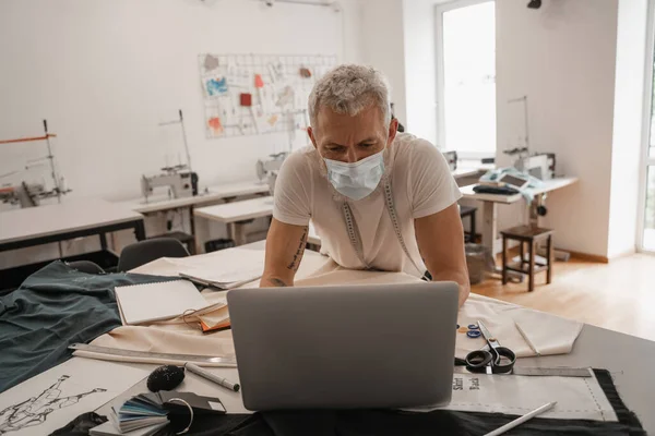 Reifer Designer in medizinischer Maske mit Laptop in Stoffnähe und Skizzen — Stockfoto