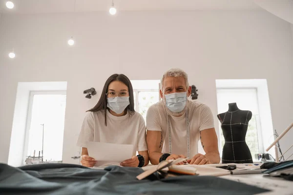 Interracial designers in medical masks looking at camera near fabric — Stock Photo