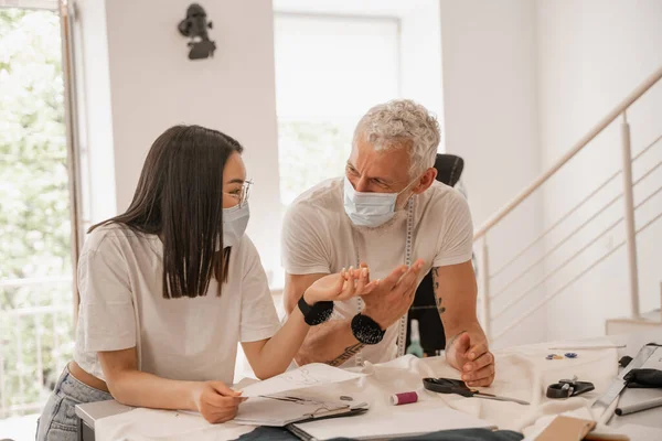 Мультиэтнические дизайнеры в медицинских масках разговаривают рядом с тканями — стоковое фото
