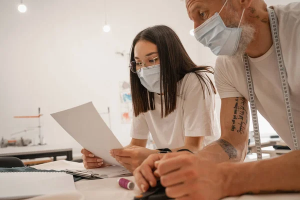 Diseñador asiático en máscara médica sosteniendo papel cerca de colega en atelier - foto de stock