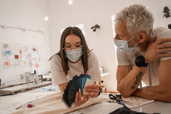 Designer asiatico in maschera medica tenendo campioni di colore vicino collega — Foto stock