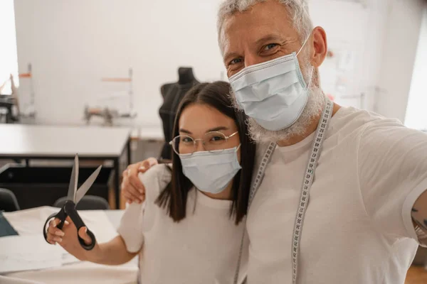 Designer in medical mask looking at camera near asian colleague with scissors — Stock Photo