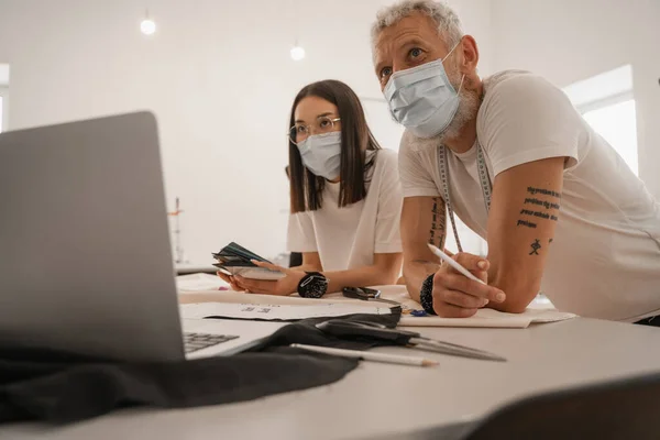 Межрасовые дизайнеры в медицинских масках работают рядом с размытым ноутбуком — стоковое фото