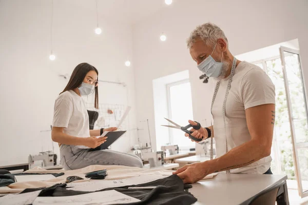 Designer in medical mask holding scissors near cloth and asian colleague — Stock Photo