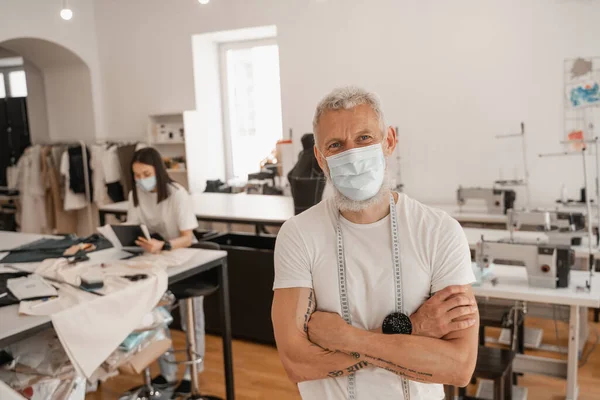 Designer mit medizinischer Maske und Maßband blickt in Atelier in die Kamera — Stockfoto