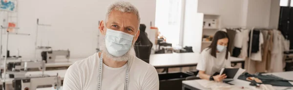 Designer en masque médical regardant la caméra près d'un collègue flou en studio, bannière — Photo de stock