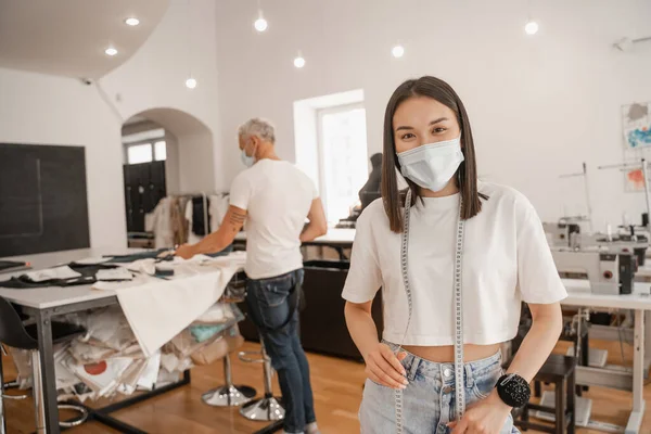 Asiatischer Designer mit medizinischer Maske und Maßband blickt in die Kamera, während sein Kollege im Studio arbeitet — Stockfoto