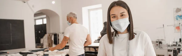 Diseñador asiático en máscara médica mirando a la cámara mientras colega borrosa trabajando en el estudio, pancarta - foto de stock