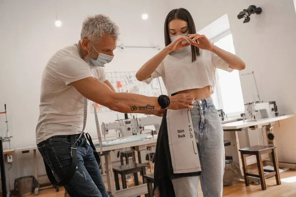 Designer in medical mask holding sewing pattern and fabric near asian customer in atelier — Stock Photo