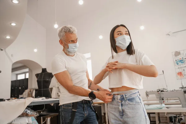 Diseñador en máscara médica midiendo cintura de cliente asiático en estudio - foto de stock