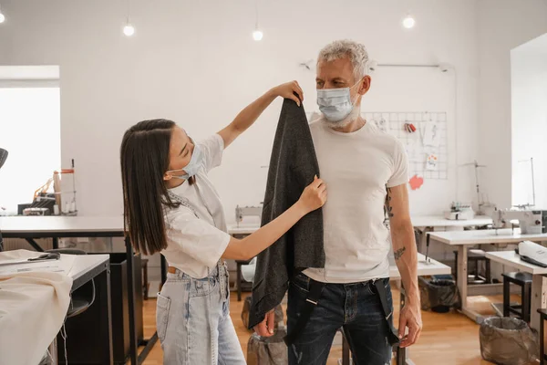 Diseñador asiático en máscara médica sosteniendo tela cerca del cliente en taller - foto de stock