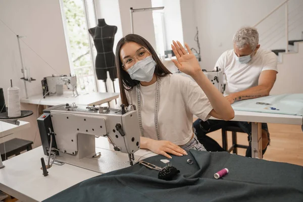 Asian seamstress in medical mask waving hand near sewing machine and colleague — Stock Photo