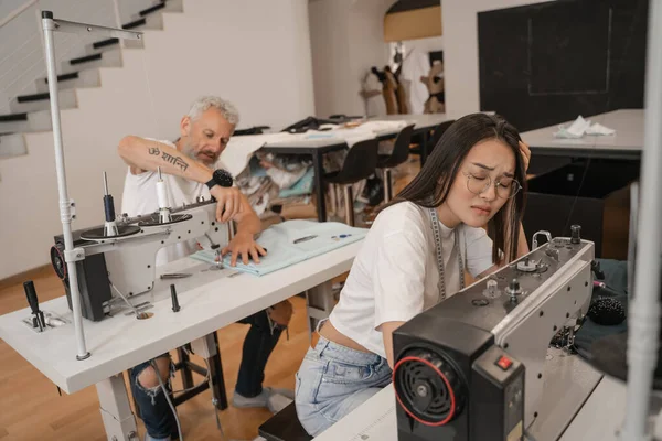 Tired asian seamstress sitting near sewing machine and colleague — Stock Photo