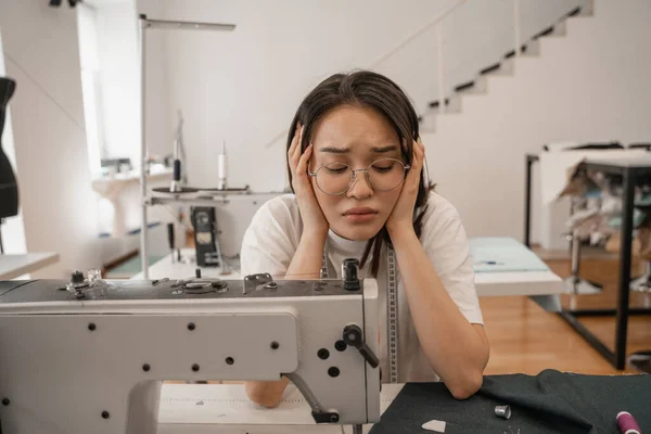 Tired asian seamstress looking at sewing machine and cloth — Stock Photo