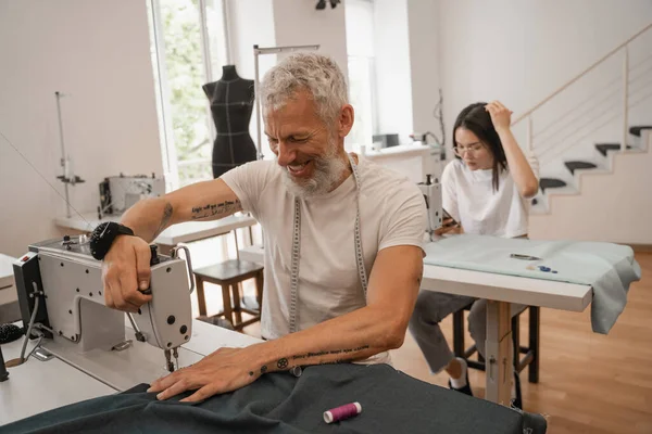 Sorrindo costura alfaiate perto colega borrado no atelier — Fotografia de Stock