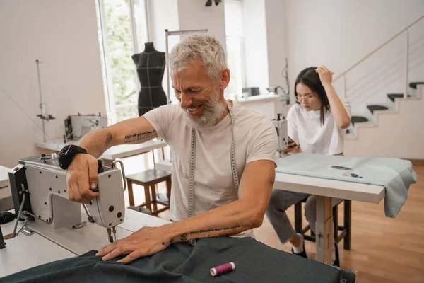 Lächelnde Schneiderin näht neben verschwommenem Kollegen im Atelier — Stockfoto