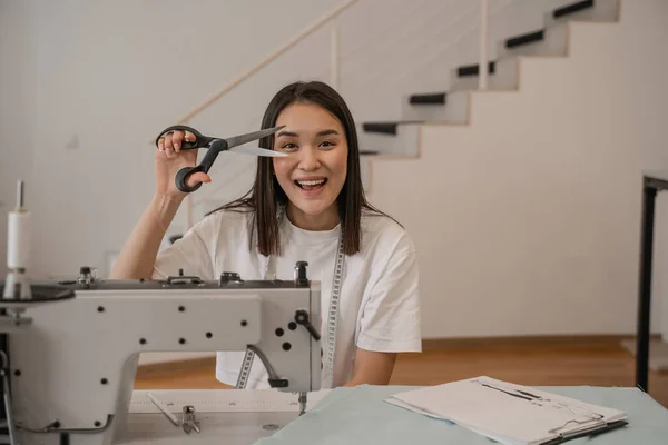 Fröhliche asiatische Designerin hält Schere in der Nähe von Nähmaschine, Stoff und Klemmbrett — Stockfoto