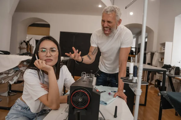 Enojado diseñador gritando cerca asiático colega en atelier - foto de stock