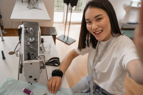 Sonriente asiático diseñador mirando a cámara cerca de máquina de coser y tela - foto de stock